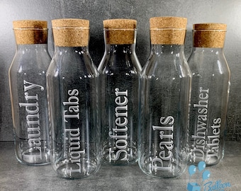 May include: Five clear glass bottles with cork stoppers and etched labels. The labels read "Laundry", "Liquid Tabs", "Softener", "Pearls", and "Dishwasher Tablets".