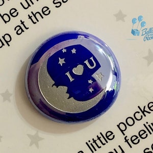 May include: A blue and silver round button with a crescent moon and stars. The moon has a silver heart with the letters "I" and "U" on either side of the heart. The text "Balloon Your Room" is visible in the background.