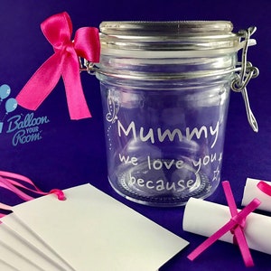 Könnte beinhalten: Ein klares Glasgefäß mit einem silbernen Deckel und einem rosa Band. Das Gefäß ist mit den Worten "Mummy we love you because" graviert. Das Gefäß ist umgeben von weißen Karten mit rosa Bändern und aufgerollten Papierstücken.