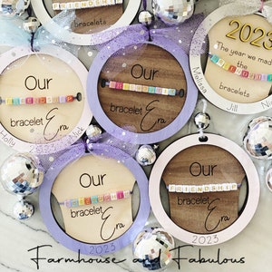 May include: Five round wooden ornaments with a purple, pink, or white painted border. Each ornament features a beaded friendship bracelet and the text "Our Friendship Bracelet Era" with the year 2023. The ornaments are decorated with silver glitter and a purple and white tulle bow.