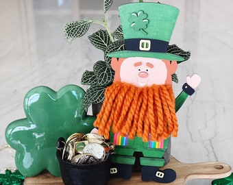May include: A green leprechaun figurine with an orange yarn beard, holding a pot of gold coins. The figurine is on a wooden round tray with a handle. There are green shamrock decorations and a gold beaded garland with shamrocks.