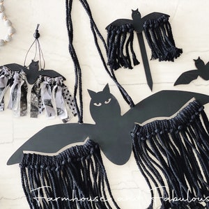May include: Four black bat-shaped wall decorations with fringe. The bats are made of cardboard and have black fringe hanging from the bottom. The bats are hanging on a white wall.