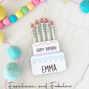 May include: A white cake card with a pink and blue frosting design. The card has the words "Happy Birthday" and "Emma" written on it. There are five dollar bills rolled up and placed on the card to look like candles.