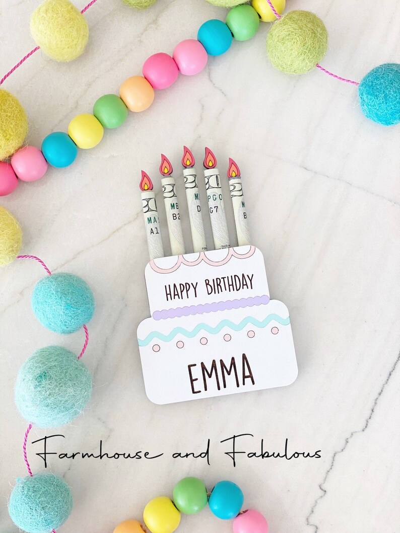 May include: A white cake card with a pink and blue frosting design. The card has the words "Happy Birthday" and "Emma" written on it. There are five dollar bills rolled up and placed on the card to look like candles.