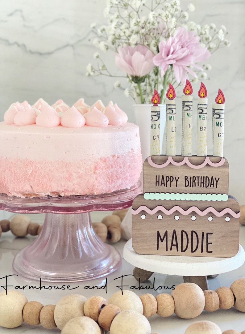 May include: A wooden cake topper with pink and green frosting, candles made of dollar bills, and the text "HAPPY BIRTHDAY MADDIE".