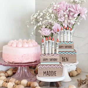May include: Two wooden cake boxes with pink and blue frosting decorations. The boxes have the words "Happy Birthday" and the names "Maddie" and "Ben". The boxes are decorated with cash money candles.