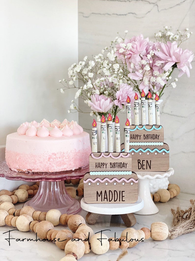 May include: Two wooden cake boxes with pink and blue frosting decorations. The boxes have the words "Happy Birthday" and the names "Maddie" and "Ben". The boxes are decorated with cash money candles.