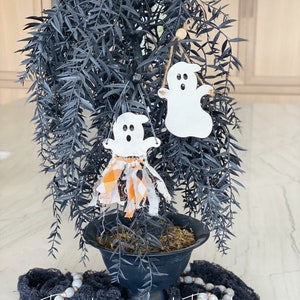 Puede incluir: Una planta en maceta negra con un soporte de metal negro. La planta tiene un adorno de fantasma blanco y negro colgando. La planta est&aacute; sentada sobre una superficie de malla negra y blanca con una guirnalda de cuentas blancas y grises.