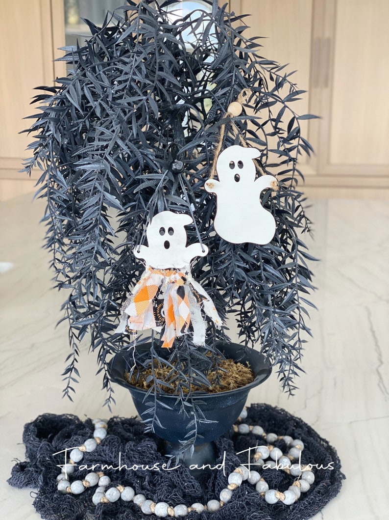 Puede incluir: Una planta en maceta negra con un soporte de metal negro. La planta tiene un adorno de fantasma blanco y negro colgando. La planta est&aacute; sentada sobre una superficie de malla negra y blanca con una guirnalda de cuentas blancas y grises.