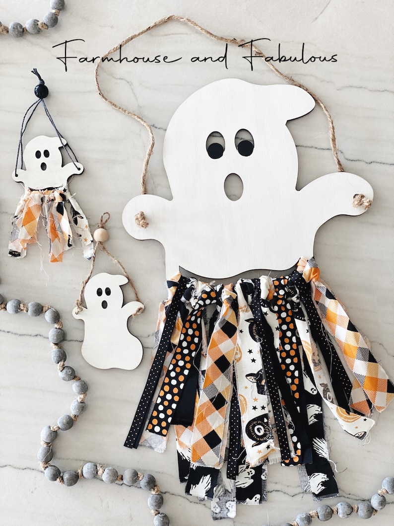 Puede incluir: Corte de fantasma de madera blanca con ojos negros y boca, colgado de una cuerda de hilo. El fantasma tiene una borla de tela negra y naranja colgando de la parte inferior. El texto "Farmhouse and Fabulous" est&aacute; escrito en negro en la parte superior de la imagen.
