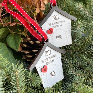 May include: Two wooden dog house ornaments with red hearts and paw prints. The ornaments are white with black trim and say "2022 The Year My Human(s) Adopted Me" with the names "Jack" and "Dug" below.