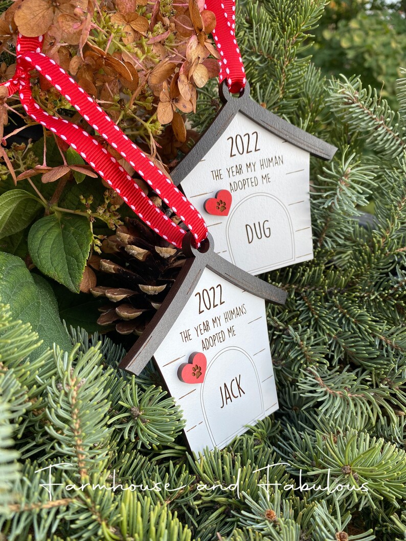 May include: Two wooden dog house ornaments with red hearts and paw prints. The ornaments are white with black trim and say "2022 The Year My Human(s) Adopted Me" with the names "Jack" and "Dug" below.