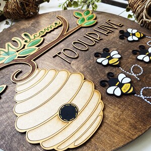 Puede incluir: Un letrero redondo de madera con el texto "Bienvenido a nuestra colmena" y una colmena con abejas volando a su alrededor. El letrero est&aacute; decorado con hojas verdes y una rama marr&oacute;n.