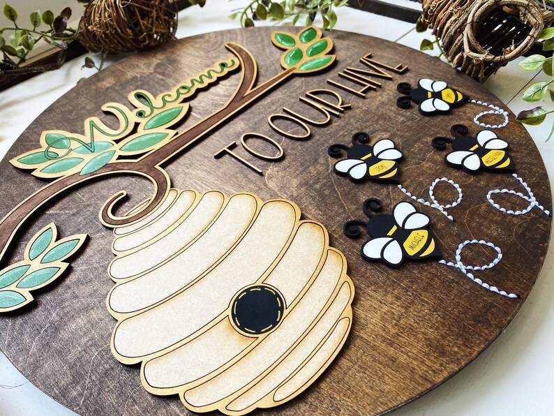 Puede incluir: Un letrero redondo de madera con el texto "Bienvenido a nuestra colmena" y una colmena con abejas volando a su alrededor. El letrero est&aacute; decorado con hojas verdes y una rama marr&oacute;n.