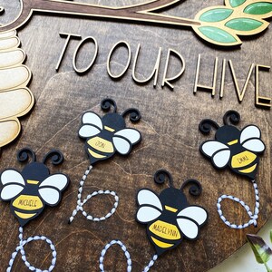 Puede incluir: Un letrero de madera con el texto "Bienvenido a nuestra colmena" y cuatro adornos con forma de abeja con los nombres "Michelle", "Jon", "Madelynn" y "Emma" escritos en ellos.