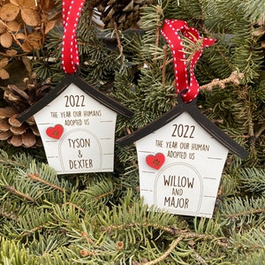 May include: Two wooden dog house ornaments with red ribbon and white polka dots. The ornaments are decorated with the year 2022, the text "The year our humans adopted us", and the names of the dogs. The first ornament says "Tyson & Dexter" and the second ornament says "Willow and Major".