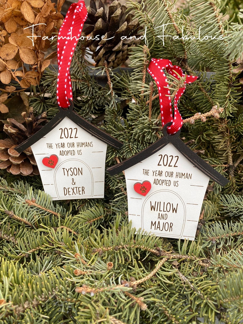 May include: Two wooden dog house ornaments with red ribbon and white polka dots. The ornaments are decorated with the year 2022, the text "The year our humans adopted us", and the names of the dogs. The first ornament says "Tyson & Dexter" and the second ornament says "Willow and Major".