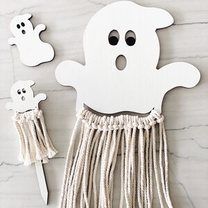 Puede incluir: Dos formas de fantasmas de madera blanca con ojos y bocas negras. El fantasma m&aacute;s grande tiene una franja de hilo blanco que cuelga de la parte inferior. El fantasma m&aacute;s peque&ntilde;o est&aacute; en un palo de madera con una franja de hilo blanco.