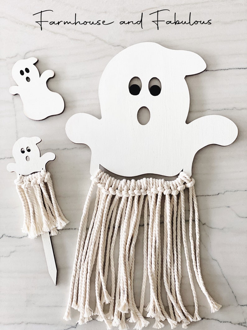 Puede incluir: Dos formas de fantasmas de madera blanca con ojos y bocas negras. El fantasma m&aacute;s grande tiene una franja de hilo blanco que cuelga de la parte inferior. El fantasma m&aacute;s peque&ntilde;o est&aacute; en un palo de madera con una franja de hilo blanco.