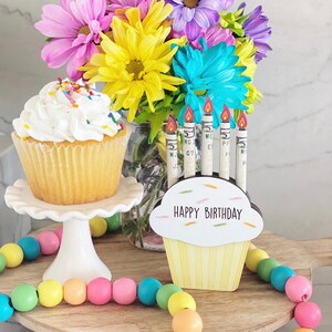 May include: A wooden cupcake stand with a yellow cupcake and the words "Happy Birthday" on it. There are six candles in the cupcake, each made from a rolled up dollar bill. The stand is decorated with a colorful beaded garland and a bouquet of flowers in a glass jar.