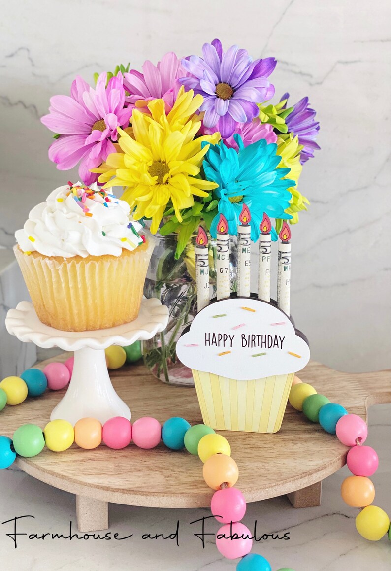 May include: A wooden cupcake stand with a yellow cupcake and the words "Happy Birthday" on it. There are six candles in the cupcake, each made from a rolled up dollar bill. The stand is decorated with a colorful beaded garland and a bouquet of flowers in a glass jar.