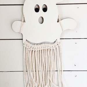 Puede incluir: Corte de fantasma de madera blanca con ojos negros y boca, colgado de una pared de macram&eacute; blanca con flecos largos.
