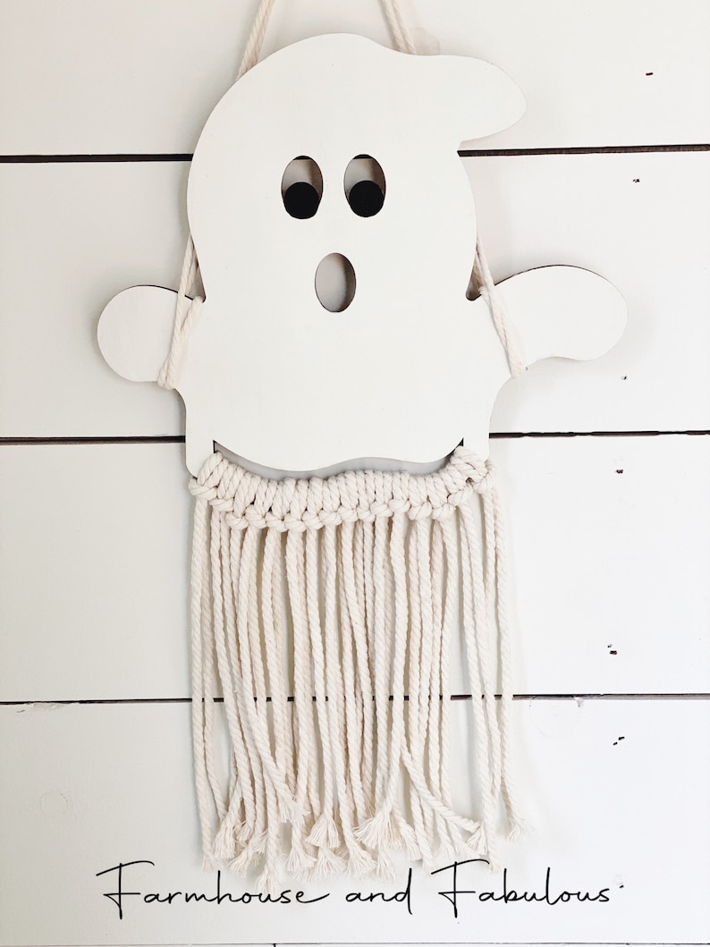 Puede incluir: Corte de fantasma de madera blanca con ojos negros y boca, colgado de una pared de macram&eacute; blanca con flecos largos.