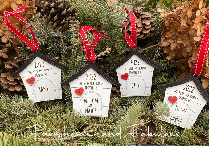 May include: Four wooden dog house ornaments with red and white ribbon. Each ornament has a year, "2022", and the text "The year our humans adopted us" or "The year my humans adopted me". The ornaments are decorated with a red heart with paw prints and the names "Tank", "Willow and Major", "Dug", and "Tyson & Dexter".