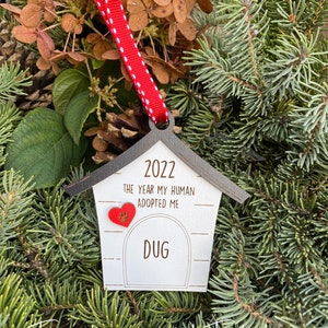 May include: White wooden dog house ornament with a red heart and the text "2022 The year my human adopted me Dug" hanging on a red and white ribbon.