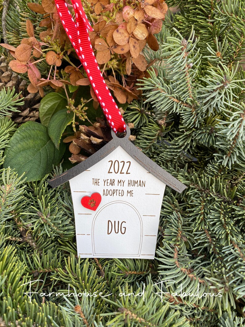 May include: White wooden dog house ornament with a red heart and the text "2022 The year my human adopted me Dug" hanging on a red and white ribbon.