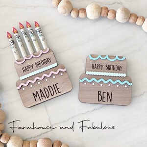 May include: Two wooden cake-shaped boxes with the words "Happy Birthday" and the names "Maddie" and "Ben" on them. The boxes are decorated with pink and blue frosting and have dollar bills sticking out of the top as candles.