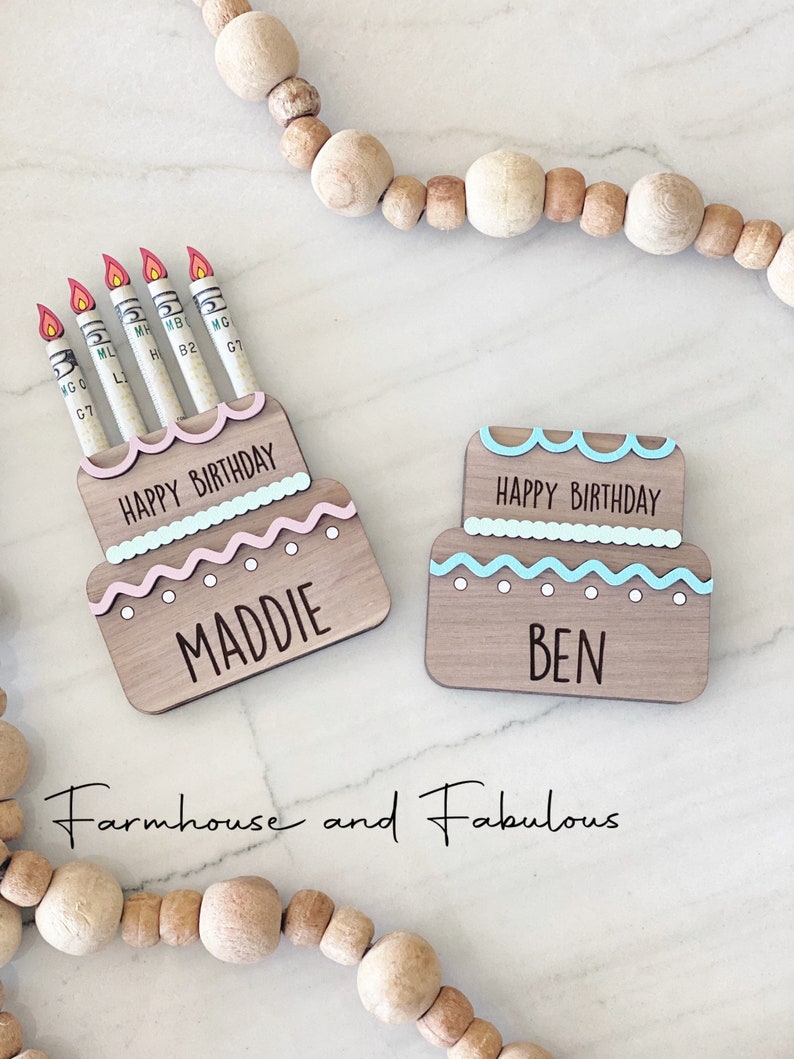 May include: Two wooden cake-shaped boxes with the words "Happy Birthday" and the names "Maddie" and "Ben" on them. The boxes are decorated with pink and blue frosting and have dollar bills sticking out of the top as candles.