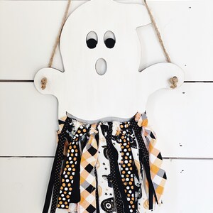 Puede incluir: Corte de fantasma de madera blanca con ojos negros y boca, colgado de una cuerda de yute. El fantasma tiene una guirnalda de tela negra, blanca y naranja unida a la parte inferior.