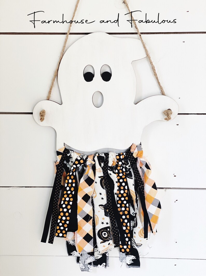 Puede incluir: Corte de fantasma de madera blanca con ojos negros y boca, colgado de una cuerda de yute. El fantasma tiene una guirnalda de tela negra, blanca y naranja unida a la parte inferior.