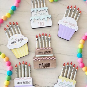 May include: Five birthday cake decorations with candles made of rolled up dollar bills. The cakes are decorated with frosting and sprinkles. The cakes are labeled with the names Lily, Maddie, and Emma. The text "Happy Birthday" is on each cake.