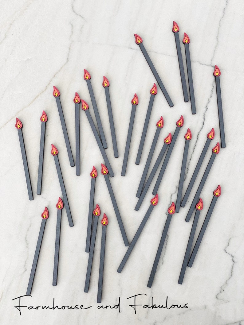 May include: A collection of 25 black wooden matchsticks with red and orange flame tips scattered on a white marble surface. The text "Farmhouse and Fabulous" is written in black cursive at the bottom of the image.