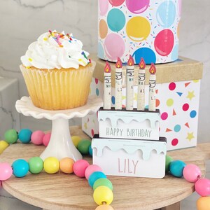 May include: A wooden birthday cake with candles and the name "Lily" on it. The cake is decorated with white frosting and colorful sprinkles. There is a cupcake with white frosting and sprinkles on a white cake stand. A colorful beaded garland is on a wooden tiered stand. The background is a white surface with a box wrapped in colorful balloons.