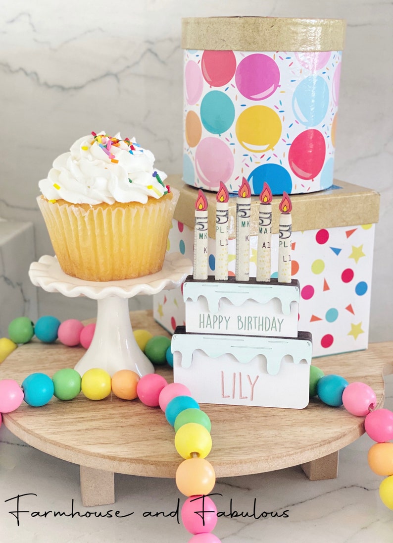 May include: A wooden birthday cake with candles and the name "Lily" on it. The cake is decorated with white frosting and colorful sprinkles. There is a cupcake with white frosting and sprinkles on a white cake stand. A colorful beaded garland is on a wooden tiered stand. The background is a white surface with a box wrapped in colorful balloons.