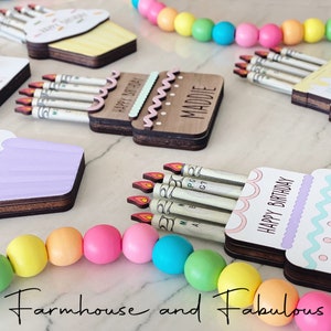 May include: A set of four wooden birthday card holders, each shaped like a cupcake or cake, with a slot for cash bills. The holders are decorated with colorful frosting, sprinkles, and the words "Happy Birthday". The names "Maddie", "Emma", and "Lily" are also visible on the holders.