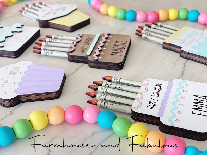 May include: A set of four wooden birthday card holders, each shaped like a cupcake or cake, with a slot for cash bills. The holders are decorated with colorful frosting, sprinkles, and the words "Happy Birthday". The names "Maddie", "Emma", and "Lily" are also visible on the holders.