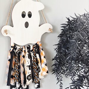 Puede incluir: Un recorte de fantasma de madera blanca con una guirnalda de tela negra y naranja colgando de &eacute;l. La guirnalda presenta varios patrones de Halloween, incluyendo lunares negros y blancos, cuadros naranja y blancos y cuadros negros y blancos. El texto "Farmhouse and Fabulous" est&aacute; escrito debajo del fantasma.