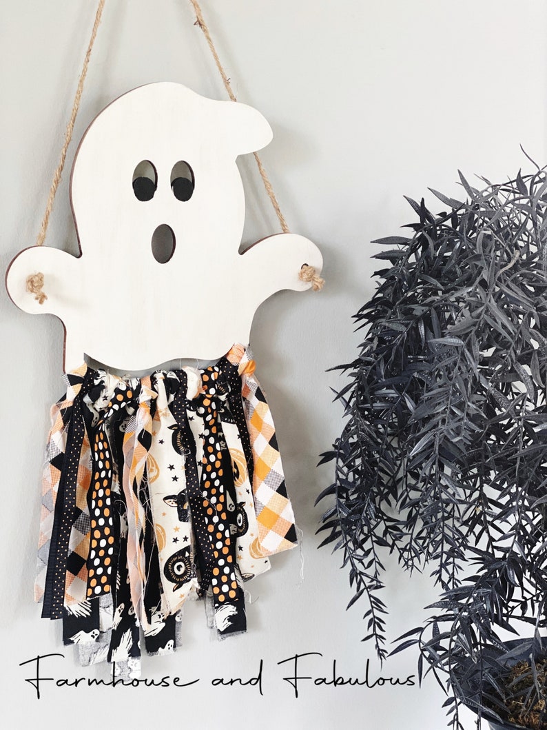 Puede incluir: Un recorte de fantasma de madera blanca con una guirnalda de tela negra y naranja colgando de &eacute;l. La guirnalda presenta varios patrones de Halloween, incluyendo lunares negros y blancos, cuadros naranja y blancos y cuadros negros y blancos. El texto "Farmhouse and Fabulous" est&aacute; escrito debajo del fantasma.