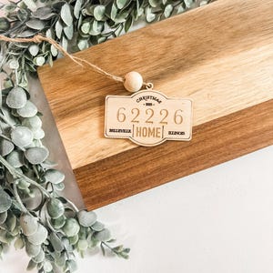 May include: A wooden Christmas ornament with the text "CHRISTMAS - 2023 - 62226 HOME BELLEVILLE ILLINOIS" hangs from a wooden bead. The ornament is on a two-tone wooden cutting board with green foliage.