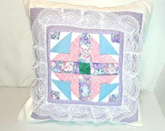 Puede incluir: Una almohada cuadrada de patchwork con un diseño floral en tonos de rosa, morado y azul. La almohada está adornada con encaje blanco y tiene un fondo blanco.