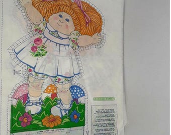 May include: A Cabbage Patch Kids fabric panel showing a doll with orange hair, a white dress with blue trim, and floral details. The doll is set against a backdrop of mushrooms and flowers. The text "LEAVE OPEN FOR STUFFING" is visible.