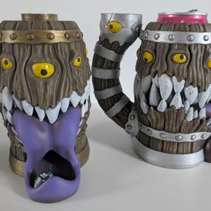 May include: Two novelty mugs with a monster design. One is a barrel-shaped mug with a purple tongue and teeth. The other is a mug with a handle and a can of soda inside. Both have yellow eyes and a wood-grain texture.