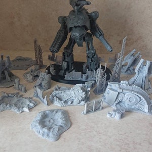 May include: A gray plastic model of a giant robot with a large gun, surrounded by smaller gray plastic models of buildings and debris. The models are for a tabletop wargame.
