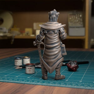 May include: A bronze painted miniature figurine of a character wearing a long robe and holding a weapon. The figurine is standing on a green cutting mat with paint pots and a dice.