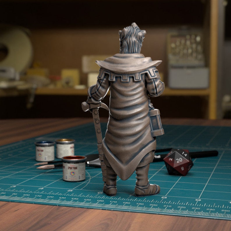 May include: A bronze painted miniature figurine of a character wearing a long robe and holding a weapon. The figurine is standing on a green cutting mat with paint pots and a dice.