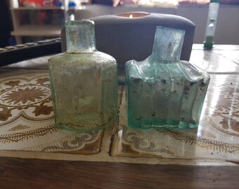 May include: Two antique glass bottles with a textured surface. The bottles are clear and have a greenish tint. They are sitting on a tiled surface.
